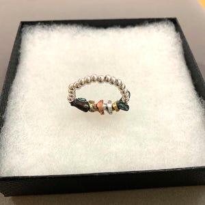 Puede incluir: Un anillo construido a partir de una serie de pequeñas cuentas esféricas plateadas pulidas. La sección inferior presenta una colección de pepitas metálicas irregulares y multicolores en tonos cobre, oro, plata y verde azulado oxidado.