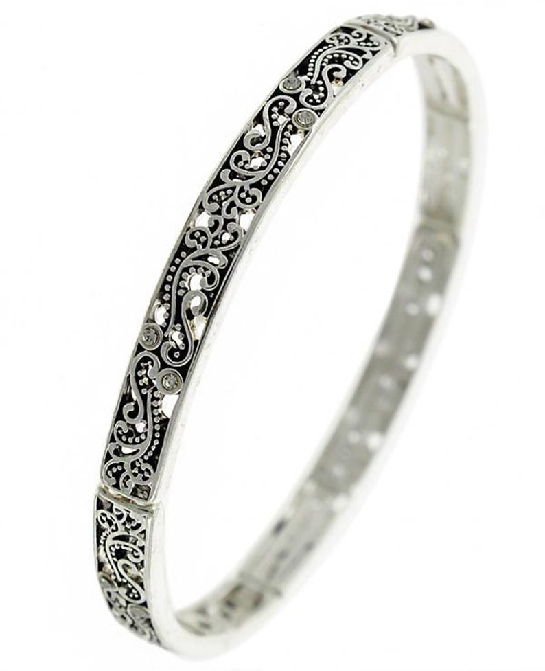 May include: A silver bangle bracelet with a delicate, intricate floral design. The design is black and features small, clear rhinestones.