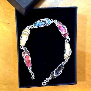 Könnte beinhalten: Ein silbernes Armband mit bunten Flip-Flop-Anhängern. Die Anhänger sind in verschiedenen Farben gehalten, darunter Rosa, Gelb und schillerndes Blau. Das Armband befindet sich in einer schwarzen Geschenkbox.