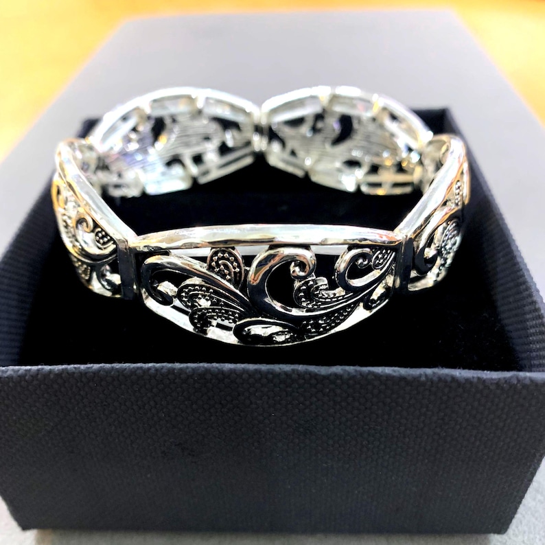 以下が含まれることがあります： 銀色のブレスレット。装飾的で透かし彫りのデザイン。ブレスレットは、渦巻く花柄とスクロールモチーフの繰り返し模様が特徴です。ブレスレットは黒い箱に入っており、ジュエリーであることを示唆しています。