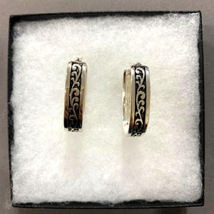 Puede incluir: Un par de pendientes de aro plateados con un dise&ntilde;o de volutas negro y ornamentado. Los pendientes rectangulares se presentan en una caja negra con acolchado blanco. Los pendientes son una joya.