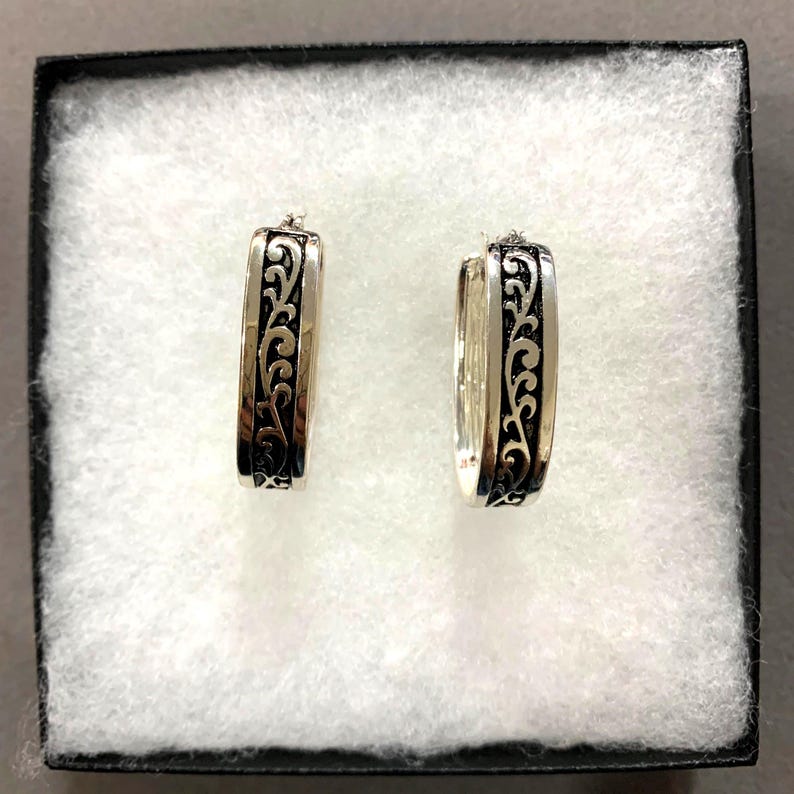 Puede incluir: Un par de pendientes de aro plateados con un dise&ntilde;o de volutas negro y ornamentado. Los pendientes rectangulares se presentan en una caja negra con acolchado blanco. Los pendientes son una joya.