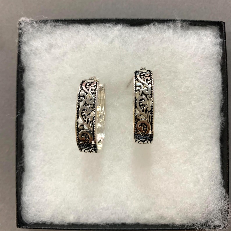 May include: A pair of silver hoop earrings with intricate, ornate scrollwork and a touch of gold-toned accents. The earrings are displayed in a black box with a white, textured interior, showcasing their detailed design.