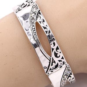 May include: Silver-toned hinged bracelet with an openwork design. The bracelet features a central oval cutout and is embellished with small, clear crystals. Intricate patterns and details adorn the metalwork.