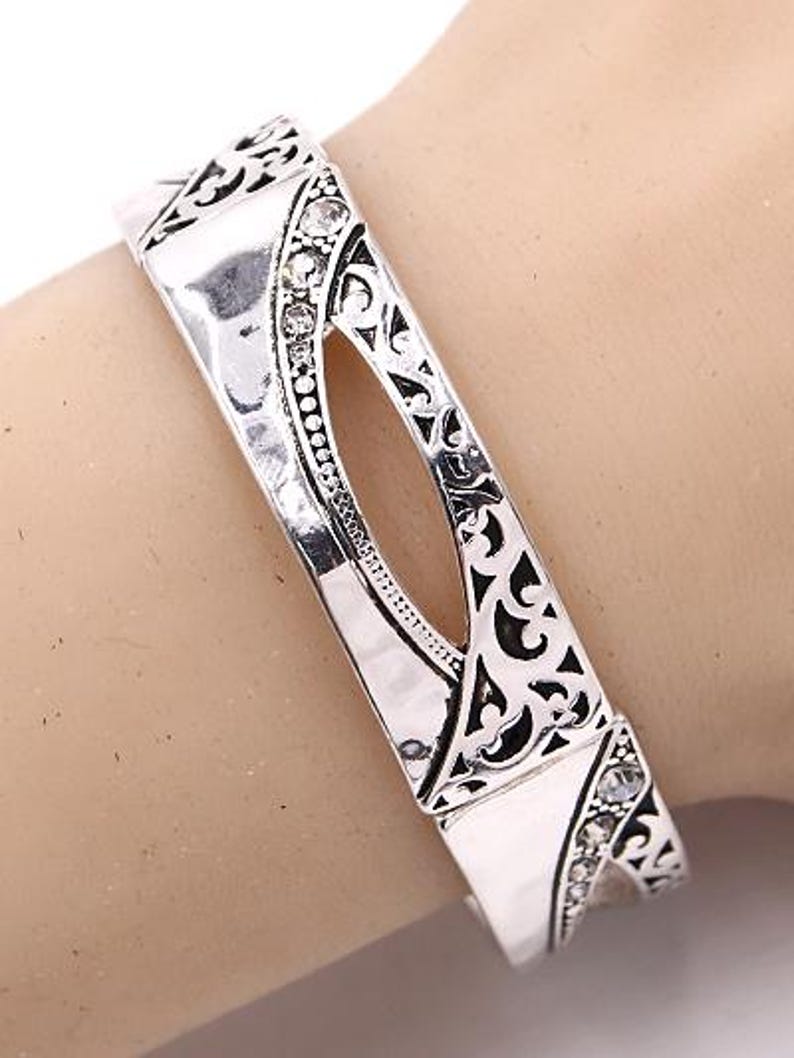 May include: Silver-toned hinged bracelet with an openwork design. The bracelet features a central oval cutout and is embellished with small, clear crystals. Intricate patterns and details adorn the metalwork.