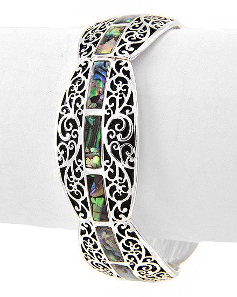 Pu&ograve; includere: Un braccialetto argentato con un design a filigrana e intarsi rettangolari in conchiglia di abalone. Il braccialetto presenta intricati motivi traforati e colori iridescenti all'interno dell'abalone. Il braccialetto &egrave; mostrato su sfondo bianco.