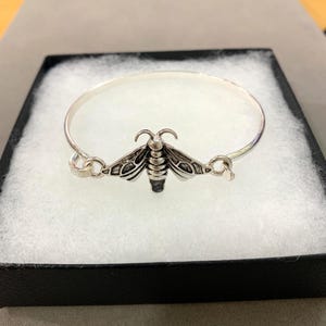 Puede incluir: Una pulsera de brazalete plateada con un dije detallado de polilla. La polilla tiene intrincados detalles de alas y cuerpo. La pulsera se exhibe en una caja negra con un interior blanco y esponjoso.