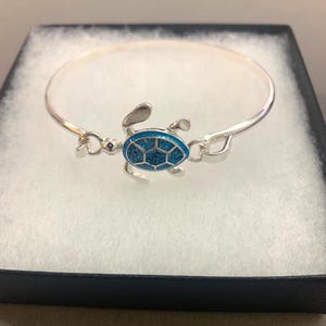 May include: A silver-toned bangle bracelet featuring a central sea turtle charm. The turtle shell is filled with bright blue, textured, sparkling material. The bracelet has a simple, thin, circular band design that connects to the turtle charm.