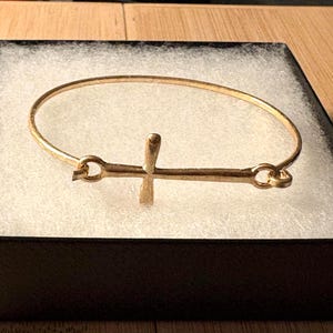 May include: A gold-colored bangle bracelet with a cross-shaped charm. The bracelet is made of a thin, circular band with a cross in the center. The cross is attached to the band with small loops. The bracelet is displayed in a black box with a white interior.