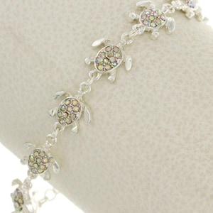 May include: A silver bracelet featuring a series of small, detailed turtle charms. Each turtle has a shell adorned with iridescent, colorful crystals. The bracelet is shown on a white surface.