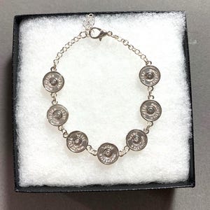 May include: Silver-toned bracelet featuring eight Winchester 38 SPL bullet casings. The bracelet is displayed in a black gift box with white padding. The chain is delicate, and the clasp is visible.