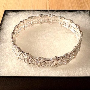 Puede incluir: Un brazalete plateado con un intrincado diseño calado, presentado en una caja negra con un interior blanco texturizado. El patrón del brazalete presenta formas entrelazadas, creando una apariencia delicada y ornamentada. La joya se exhibe sobre una superficie clara.
