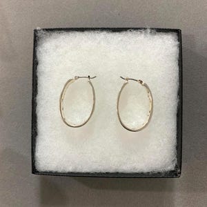 Puede incluir: Un par de pendientes de aro ovalados plateados presentados en una caja de joyer&iacute;a negra con acolchado blanco. Los pendientes tienen un dise&ntilde;o simple y elegante, con un patr&oacute;n texturizado a lo largo del borde exterior. Los pendientes se muestran sobre un fondo neutro.