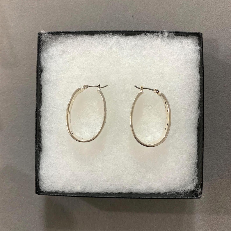 Puede incluir: Un par de pendientes de aro ovalados plateados presentados en una caja de joyer&iacute;a negra con acolchado blanco. Los pendientes tienen un dise&ntilde;o simple y elegante, con un patr&oacute;n texturizado a lo largo del borde exterior. Los pendientes se muestran sobre un fondo neutro.