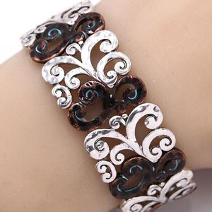 Może przedstawiać: Bransoletka srebrna i miedziana z kręconym wzorem kwiatowym. Bransoletka jest wykonana z metalu i ma fakturowane wykończenie.