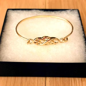 May include: A gold-colored bangle bracelet with an intricate Celtic knot design at the center. The bracelet is presented in a black box with a white interior, creating a striking contrast. The bracelet's design is a continuous loop.