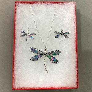 May include: A silver-toned jewelry set consisting of a necklace and a pair of drop earrings. Each piece features a dragonfly design with iridescent, multi-colored inlay wings and a polished silver body, presented in a red-bordered display box.