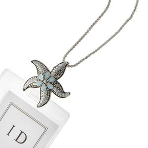 May include: A silver-toned necklace featuring a starfish pendant. The starfish has a light blue and silver design with a detailed pattern. The necklace is on a white background with a clear plastic ID holder in the lower left corner.