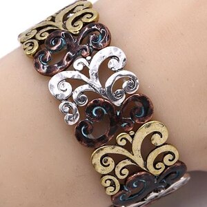 Może przedstawiać: Bransoletka srebrna, miedziana i złota z ozdobnym, wirującym wzorem. Bransoletka jest wykonana z metalu i ma fakturowane wykończenie.