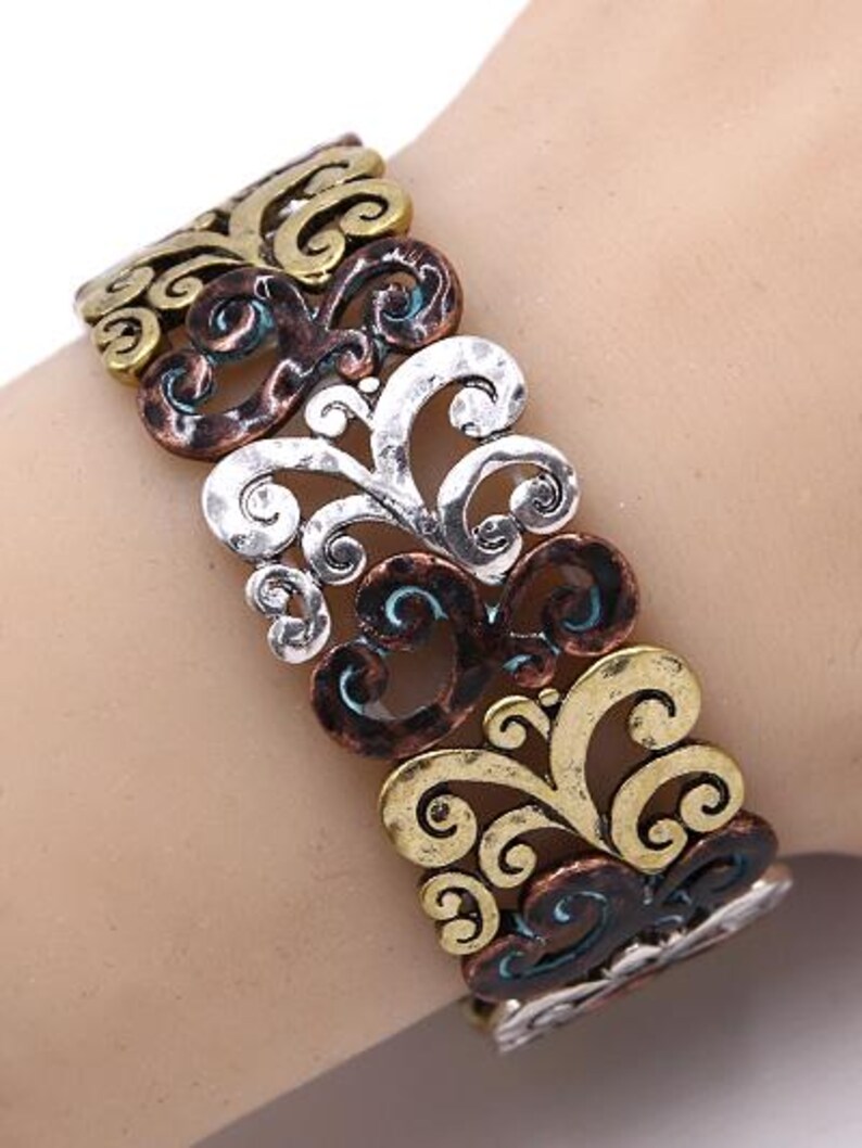 Może przedstawiać: Bransoletka srebrna, miedziana i złota z ozdobnym, wirującym wzorem. Bransoletka jest wykonana z metalu i ma fakturowane wykończenie.