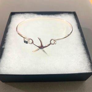 May include: A silver-toned bangle bracelet featuring a central starfish charm. The bracelet has a smooth, polished finish and a circular band design. The starfish charm is attached to the band with small metal loops on either side.