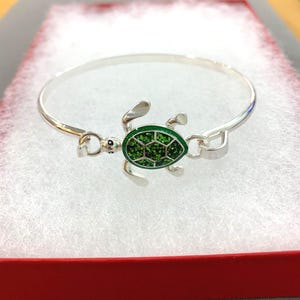 Può includere: Un braccialetto a cerchio d'argento con un ciondolo a forma di tartaruga. La tartaruga ha un guscio verde strutturato e arti argentati. Il braccialetto è presentato in una scatola rossa con imbottitura bianca.