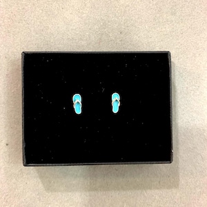 Puede incluir: Un par de pendientes de botón con forma de chanclas turquesas y doradas, presentados en una caja negra. Los pendientes son pequeños y detallados, con una base turquesa brillante y correas doradas. La caja es rectangular.