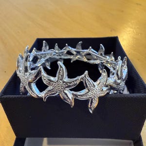 Könnte beinhalten: Ein silberfarbener Seestern-Kopfschmuck, präsentiert in einer schwarzen Schachtel. Der Kopfschmuck weist mehrere Seestern-Designs auf, die durch ein zartes Band verbunden sind. Die Seesterne haben strukturierte Oberflächen, und das Gesamtdesign ist aufwendig und detailliert. Es ist ein dekoratives Accessoire.