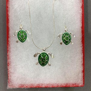 May include: A silver-toned jewelry set featuring a turtle design. The set includes a necklace with a pendant and a pair of earrings. Each piece has a green, gem-filled turtle shell and is presented in a red box with white padding.