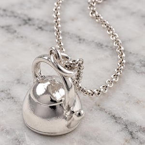 May include: A polished silver-toned pendant shaped like a miniature tea kettle, featuring a rounded body, a curved handle, and a small spout. The pendant is suspended from a matching silver-toned link chain, creating a cohesive metallic accessory.