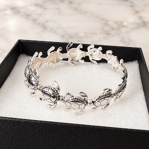 May include: A silver-toned bracelet composed of a series of connected sea turtle charms. Each turtle features detailed shell markings and is linked together to form a circular band. The bracelet is presented inside a black box with a white interior.