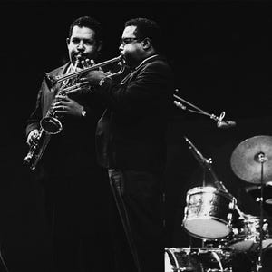 Może przedstawiać: Czarno-białe zdjęcie zespołu jazzowego występującego na scenie. Dwóch muzyków jest na pierwszym planie, jeden gra na saksofonie, a drugi na trąbce. Widoczny jest również mikrofon i zestaw perkusyjny.