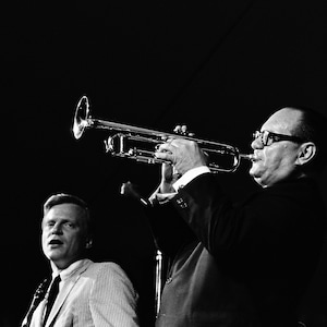 May include: A man plays a trumpet while another man stands beside him. The man playing the trumpet is wearing glasses and a suit. The man standing beside him is wearing a light-coloured shirt and tie.