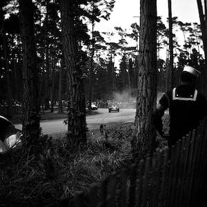 May include: A black and white photo of a man in a sailor's uniform watching a race car go by on a track through a wooded area.