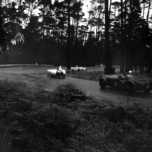 May include: Three vintage race cars driving on a winding road through a forest. The cars are black and white and have numbers on the side. The road is paved and the forest is dense with trees.