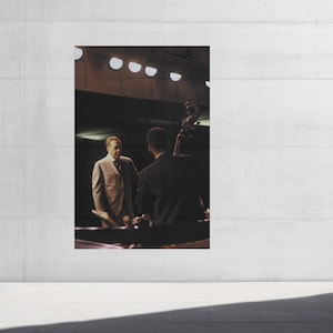 May include: A large framed print of a jazz performance. The image shows two men in suits, one standing and one playing a double bass. The print is mounted on a concrete wall, with a person standing in front of it.