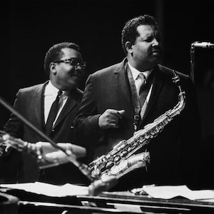 Może przedstawiać: Dwóch mężczyzn w garniturach, jeden gra na saksofonie, drugi patrzy. Saksofon jest srebrny i ma czarną ustnik. Mężczyźni są w słabo oświetlonym pomieszczeniu, a tło jest rozmyte.