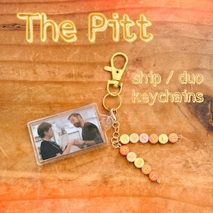 May include: A rectangular clear acrylic keychain featuring a photograph of two people holding hands, attached to a gold-toned lobster clasp. Two strands of colourful letter beads spell out the names HUCKLE and ROBBY in yellow and orange tones.