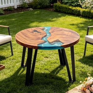 May include: A round table featuring a natural wood grain surface with a central river-like inlay of translucent blue resin. The table is supported by four black metal legs and is positioned outdoors on a green lawn between two chairs.