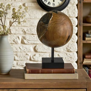 May include: A decorative wooden sphere with a dark brown finish, mounted on a black metal stand and a square base. The sphere sits atop two leather-bound books and a light-colored book. A gray vase with greenery is to the left.