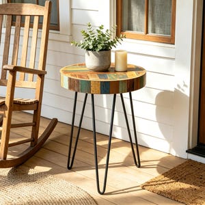 May include: A round side table with a multicolored reclaimed wood top and three black metal hairpin legs. A gray ceramic pot containing a green plant and a cylindrical cream-colored candle are placed on the surface of the wooden table.