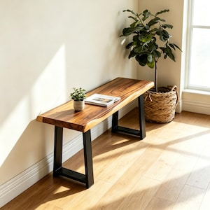 May include: A rectangular wooden bench featuring a natural live edge top with visible wood grain patterns in shades of brown. The bench is supported by two black metal U-shaped legs. A small potted green plant and an open book rest on the surface.