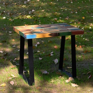 May include: A small, rectangular table with a colorful, patchwork top made of reclaimed wood. The table has black metal legs with a simple, A-frame design. The table is sitting on a grassy surface with fallen leaves.