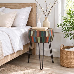 May include: A round side table featuring a drum-shaped top constructed from vertical wooden slats in various colors including blue, green, orange, and brown, supported by three thin black metal hairpin legs standing on a light wood floor.