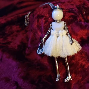 May include: A silver doll pendant necklace. The doll features a white ball head, a white dress with tassels, and silver arms and legs. It is holding a small purse. The necklace is on a silver chain.