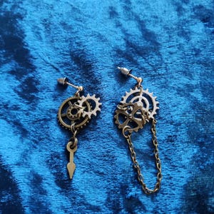 May include: Steampunk earrings with bronze-coloured gear designs. One earring has a small key pendant, the other a chain. The earrings are designed for pierced ears. The gears are approximately 2-3 cm in diameter.