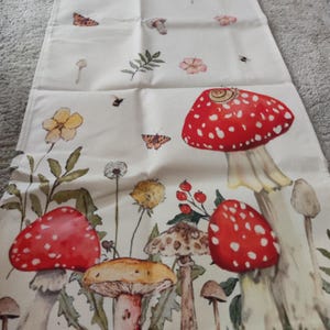 Könnte beinhalten: Cremefarbener Tischläufer mit einem skurrilen botanischen Design. Der Läufer ist mit Illustrationen von rot-weiß gepunkteten Pilzen, Blumen, Schmetterlingen und anderen Naturelementen verziert. Das Design ist auf einem weichen, strukturierten Stoff gedruckt.