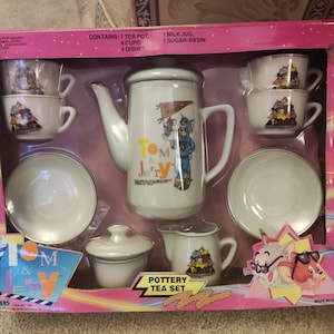 May include: A white Tom & Jerry pottery tea set in a pink box. The set includes a teapot, cups, saucers, a sugar basin, and a milk jug. The teapot and cups feature Tom & Jerry graphics. The box reads "Pottery Tea Set".