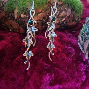 May include: A pair of silver-coloured drop earrings. Each earring has a green gemstone at the top and a series of detailed leaf charms. The leaves are arranged along a slender, curved bar, creating an elegant, nature-inspired design.