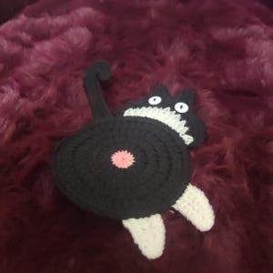 May include: A handmade crochet cat coaster, featuring a black body with a pink center, white paws, and a white and black head with white eyes. The cat's tail is curved upwards. The coaster is designed to protect surfaces from drink rings.
